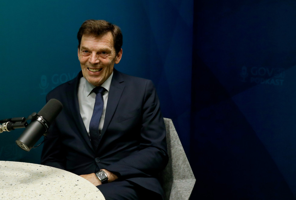 Moški srednjih let do starejši, v temnem poslovnem oblečnem suknjiču, srajci in kravati, sedi nasmejan pred mikrofonom v studiu z modro ozadjem. Ozadje vsebuje logotip »GOVSI PODKAST«. Moški ima kratke temne lase z nekaj sivine in nosi uro na levi roki. P