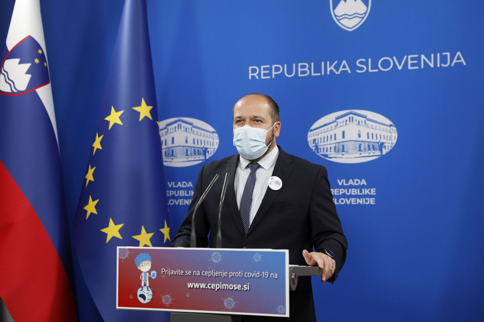 Minister za zdravje Janez Poklukar stoji za govorniškim pultom, ob strani na levi zastava, v ozadju na steni emblemi slovenske vlade.