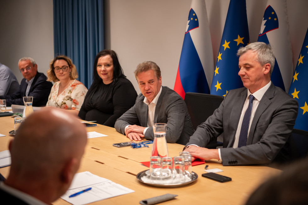 Predsednik vlade za mizo govori. Poleg njega sedijo moški in ženske. Zadaj slovenska in evropska zastava.