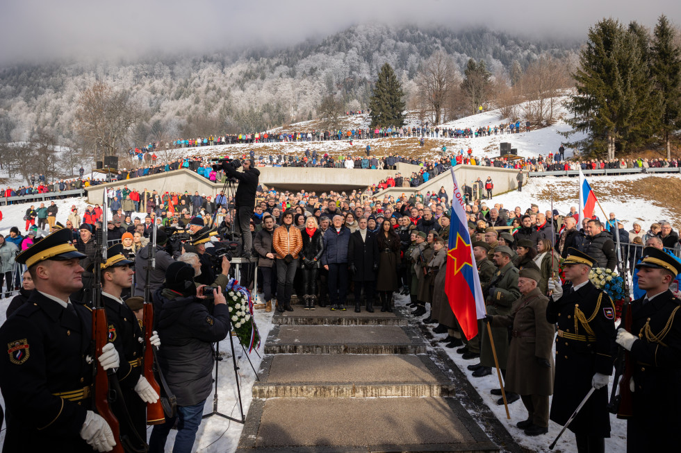 Predsednik vlade in mnogo drugih stojijo zunaj na prireditvi. Pred njimi častna straža in zastave.