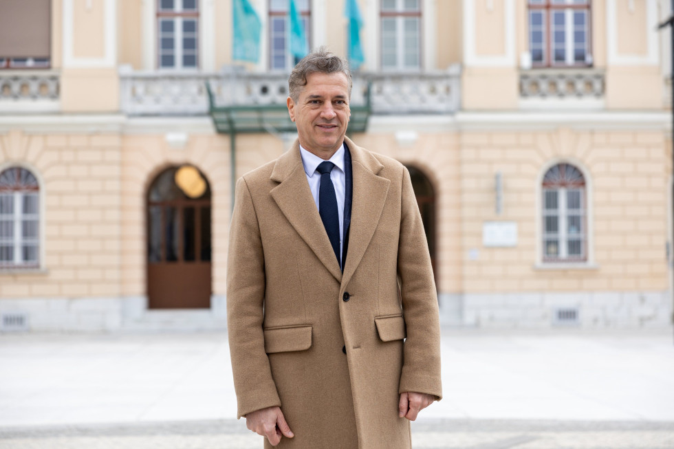 Predsednik vlade, oblečen v plašč, stoji pred stavbo in se smehlja.