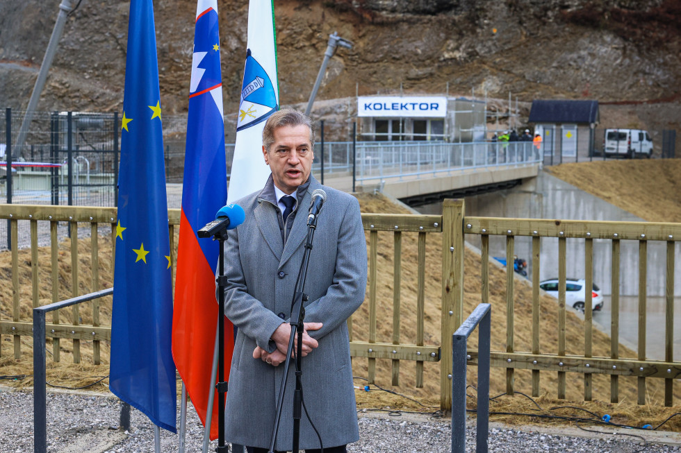 Robert Golob stoji, na levi so zastave, za njim je most.