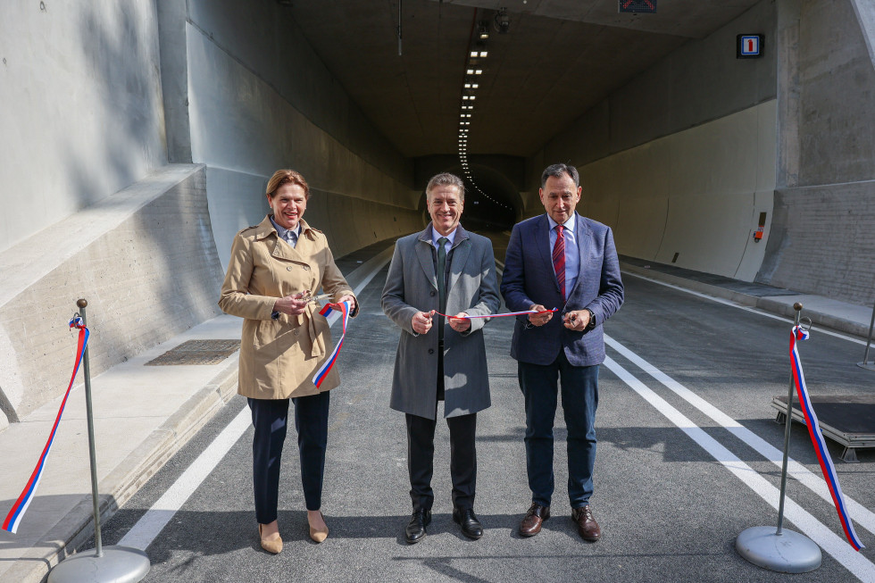 Robert Golob, Alenka Bratušek in Andrej Ribič stojijo na cesti pred tunelom, v rokah držijo škarje in trakove.