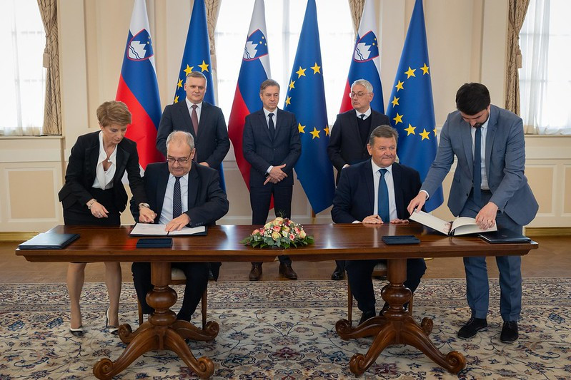 Podpis uradnega dokumenta ob prisotnosti slovenskih in evropskih predstavnikov, v ozadju zastave EU in Slovenije.