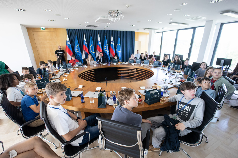 Otroci sedijo v sejni dvorani Vlade Republike Slovenije in poslušajo predstavitev.