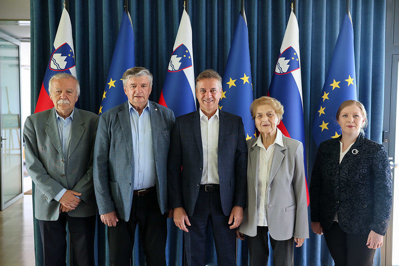 Pet oseb stoji pred zaveso, okrašeno z zastavami Republike Slovenije in Evropske unije. Trije moški in dve ženski pozirajo za skupinsko fotografijo. Vsi so formalno oblečeni, v središču stoji moški v modrem suknjiču, ki se smehlja.
