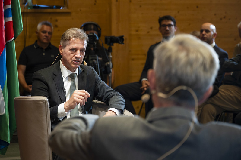 Moški v obleki govori med javnim pogovorom, opremljen z naglavnim mikrofonom. V ospredju je neizostren sogovornik, v ozadju pa poslušalci in snemalna ekipa.