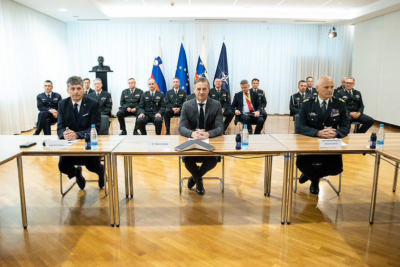 Predsednik vlade in še dva moška sedita za mizo. Zadaj več moških v vojaških uniformah sedi na stolih. Zadaj zastave. 