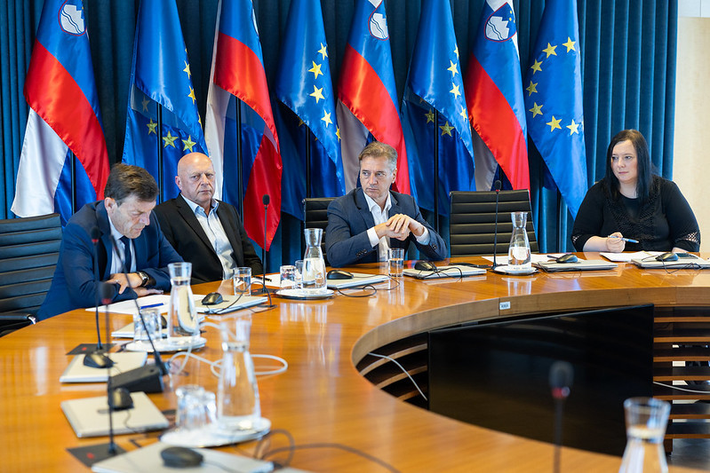 Ljudje sedijo v ospredju okrogle mize. V ozadju so razporejene zastave Slovenije in Evropske unije. Vsi zbrano spremljajo razpravo in imajo pred seboj dokumente in mikrofone.