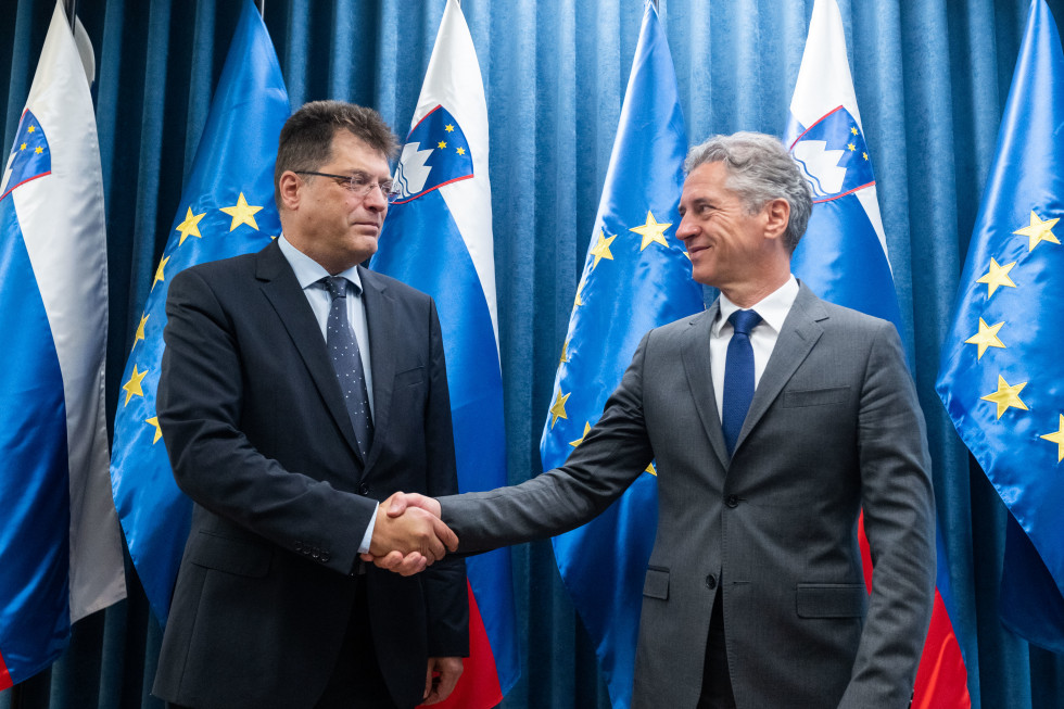 Janez Lenarčič  in Robert Golob se rokujeta, v ozadju so zastave.