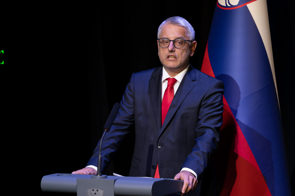 minister ob govorniškem pultu, v ozadju slovenska zastava