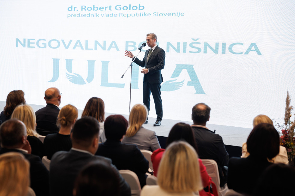 Robert Golob stoji na odru za mikrofonom, v ospredju so udeleženci otvoritve bolnišnice. 