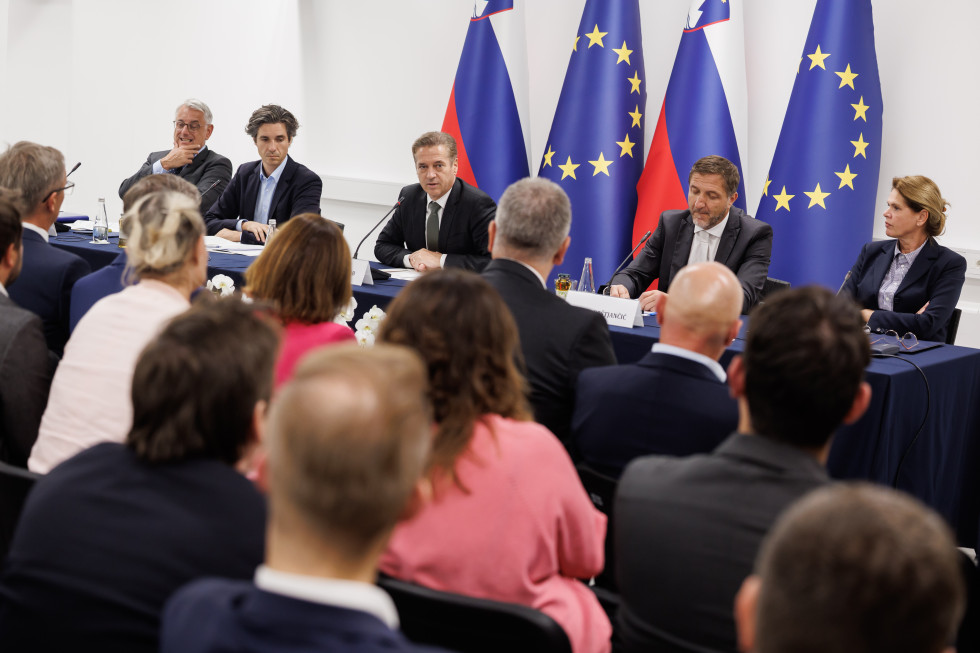 Robert Golob in drugi udeleženci srečanja sedijo, v ozadju so zastave.