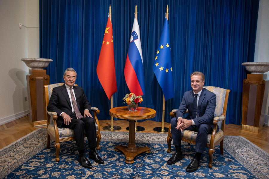 The two men are sitting opposite each other, with a small round table between them. In the background, there are flags.