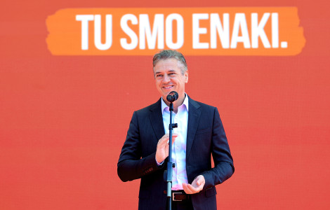PV Tu smo enaki (The prime minister stands behind the microphone and is smiling. In the background, there is a sign that says We are equal here.)