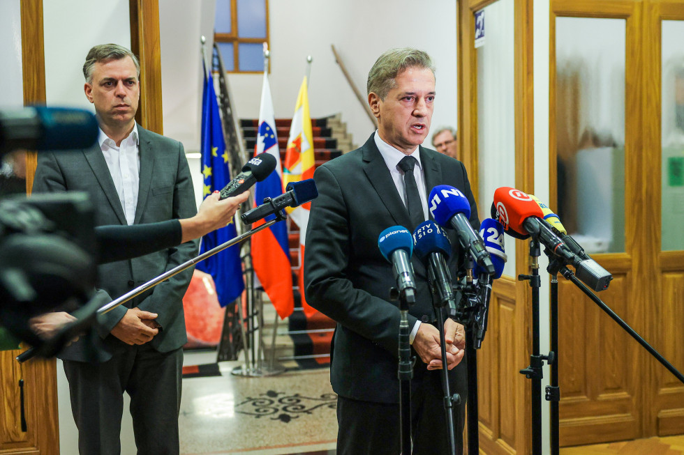 Gregor Macedoni in Robert Golob stojita, pred njima so mikrofoni, v ozadju so zastave.