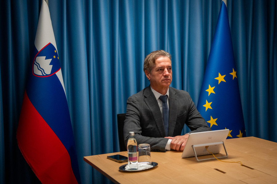 Robert Golob is sitting at a table with a laptop in front of him, a bottle of water and a glass on his left. There are flags on the left and right, and a blue curtain in the background.