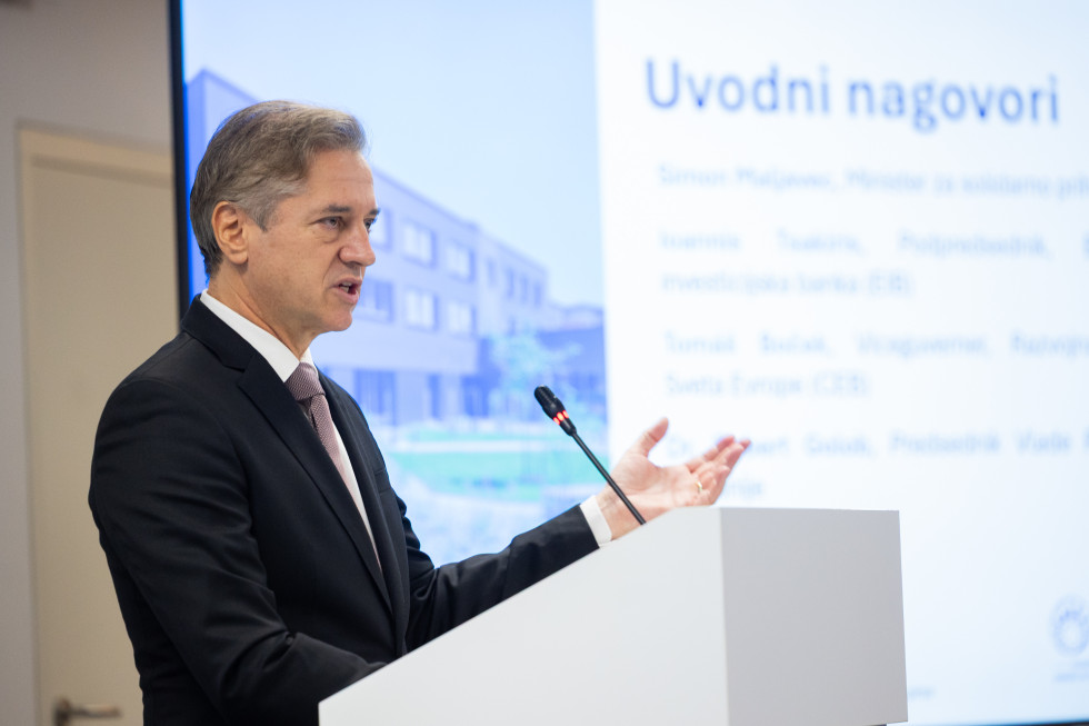 Robert Golob stoji za govornico, pred njim je mikrofon, v ozadju so napisani napovedani govorci konference. 