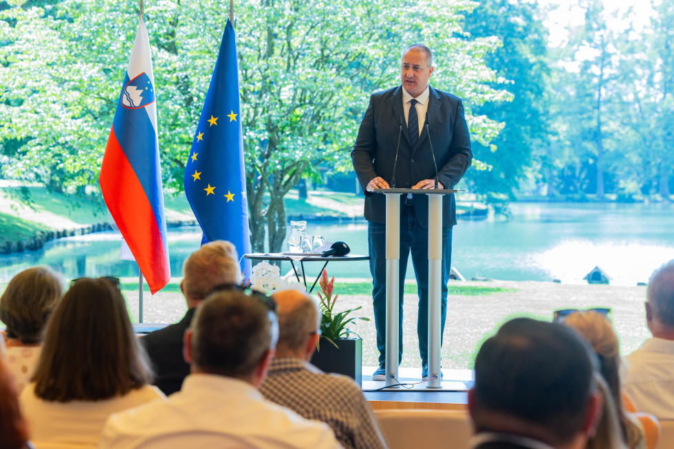 Državni sekretar stoji za govornico in nagovarja udeležence. V ozadju so drevesa in jezero.