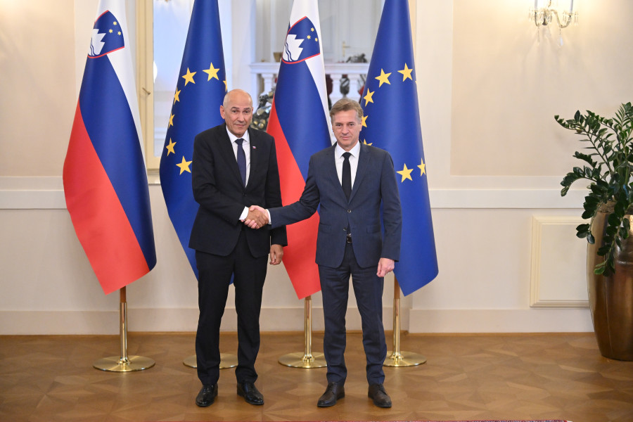 Dve osebi stojita v notranjem prostoru in pozirata za fotografijo. V ozadju so zastave Slovenije in Evropske unije.