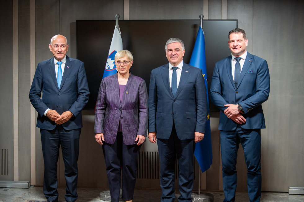 Štiri osebe stojijo pred velikim zaslonom, za njimi slovenska in evropska zastava.