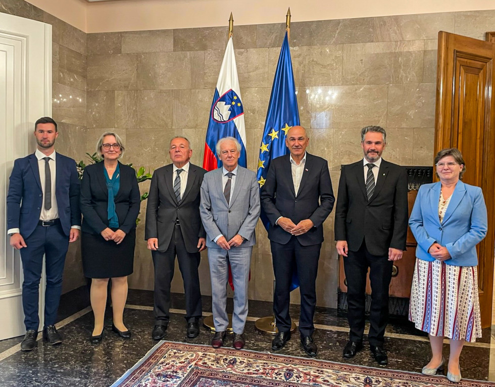 Člani Fiskalnega sveta in predstavniki Ministrstva za finance stojijo, Janez Janša je na sredini, v ozadju so zastave.