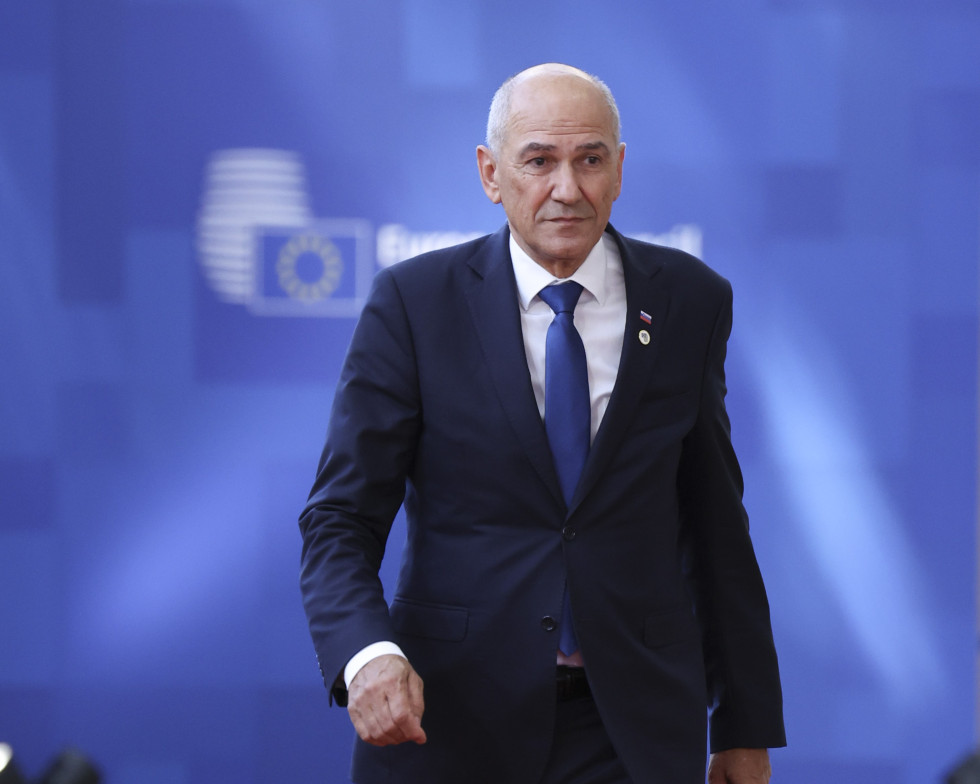 Moški v temni obleki in modri kravati hodi po prizorišču vrha Evropske unije, v ozadju je moder pano z zabrisanim logotipom Evropskega sveta.