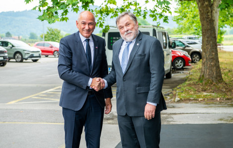 PV2 2134 (Two men in dark suits shake hands and smile in a parking lot under leafy trees, with hills in the background.)