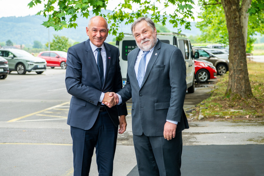Two men in dark suits shake hands and smile in a parking lot under leafy trees, with hills in the background.