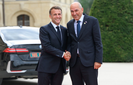 Pariz (Two men in dark suits shake hands and smile outdoors, with a black car and a stone building in the background.)