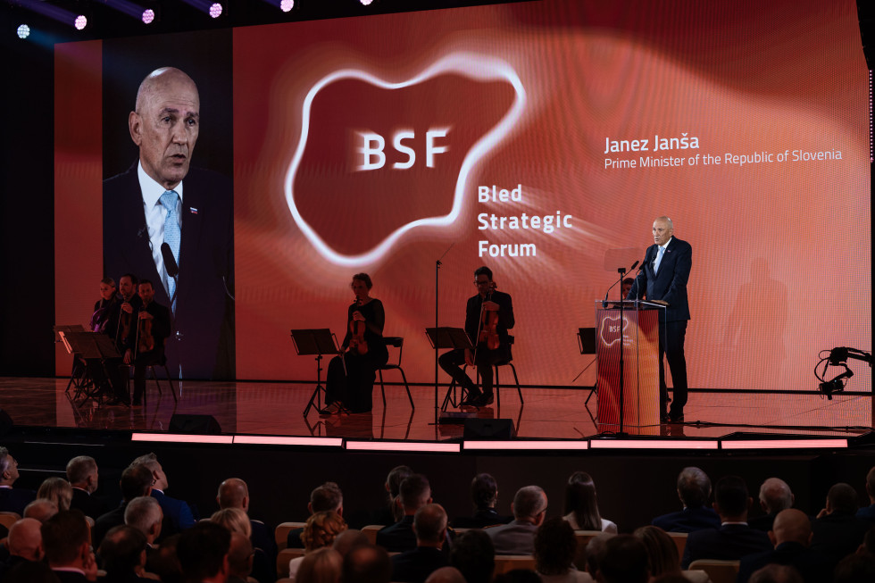 Janez Janša is standing on the right behind the lectern; two musicians are seated in the centre; and on the right is a screen showing an image of Janez Janša. In the background, in the centre, are the symbols of the Bled Strategic Forum.
