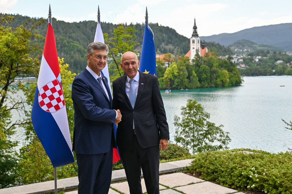 Andrej Plenković in Janez Janša stojita in se rokujeta, v ozadju na levi so zastave, na desni Blejsko jezero z Blejskim otokom.