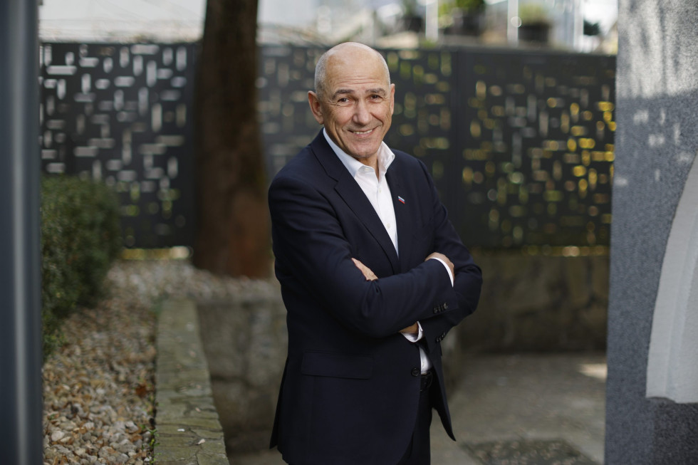 Janez Janša stoji s prekrižanimi rokami, v ozadju je betonska ograja in drevo.