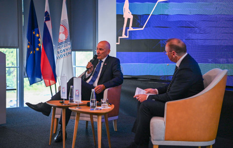 PV Forum obrti in podjetništva 1 02092026 (Janez Janša is sitting; to his right sits the moderator of the discussion; in front of them are small tables; to the left is a window, and in front of it are flags.)
