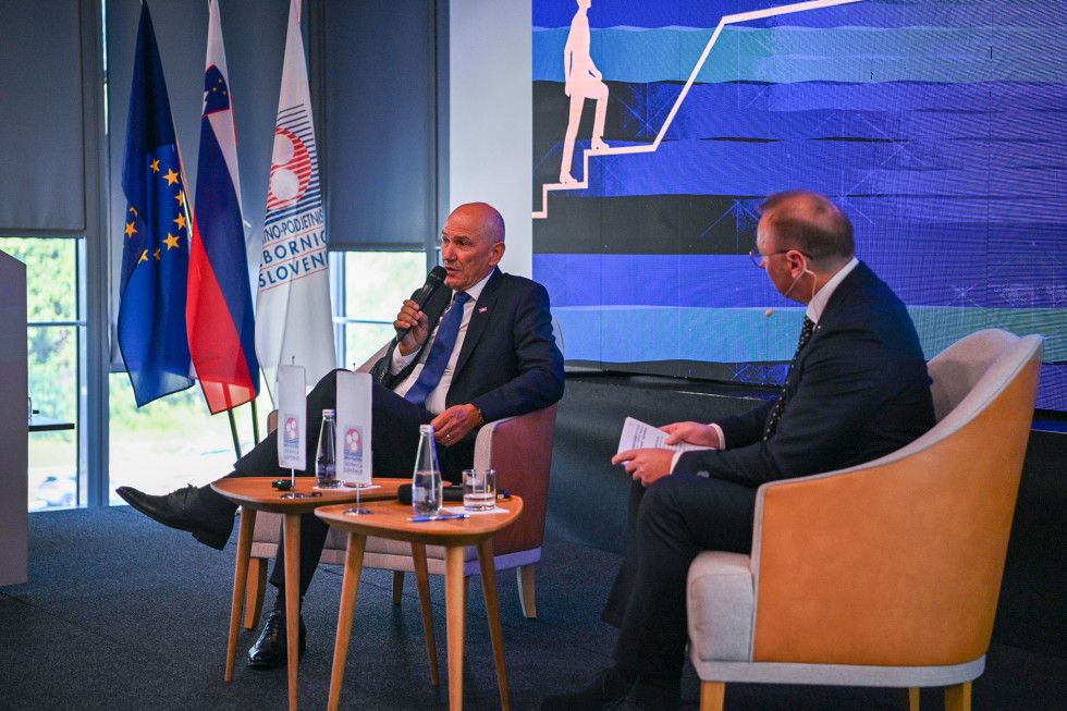 Janez Janša is sitting; to his right sits the moderator of the discussion; in front of them are small tables; to the left is a window, and in front of it are flags.