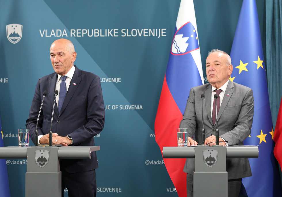 Dva moška stojita za govornicama, za njima je slovenska zastava in modro ozadje z napisom Vlada Republike Slovenije