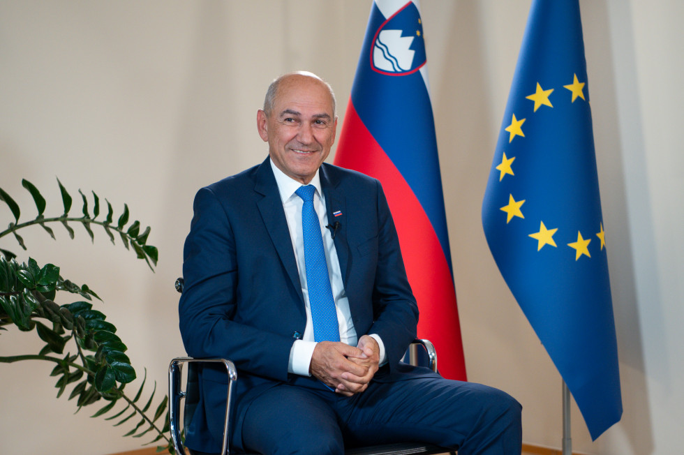 Janez Janša is sitting; in the background, on the left, is a plant, and on the right are flags.