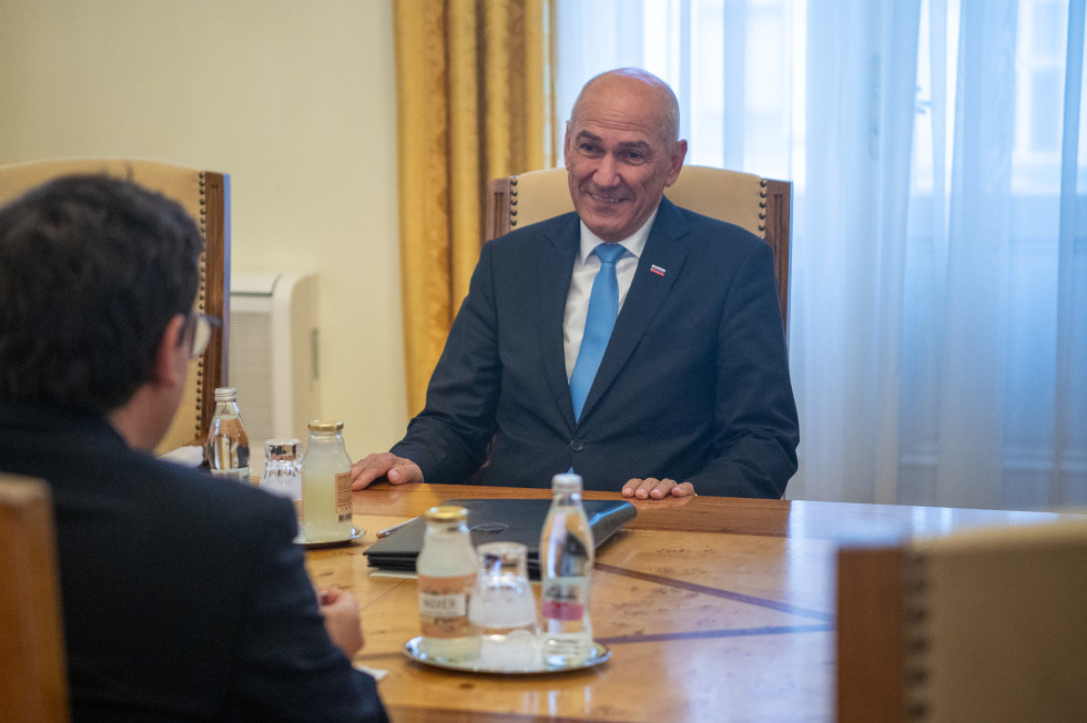 Moški v suknjiču in kravati sedi za leseno mizo, na kateri je pijača, in se smeji nasproti sedečemu moškemu