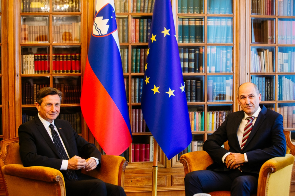Borut Pahor in Janez Janša sedita v foteljih, med njima stojijo zastave, v ozadju je vitrina.