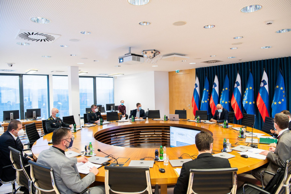 Janez Janša in prisotni sedijo za okroglo mizo v vladni konferenčni sobi. V ozadju so okna in zastave.