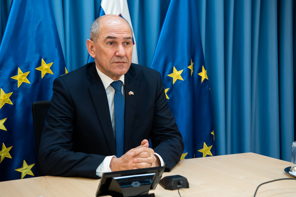 Predsednik vlade Janez Janša sedi za mizo, v ozadju so zavesa in zastave.