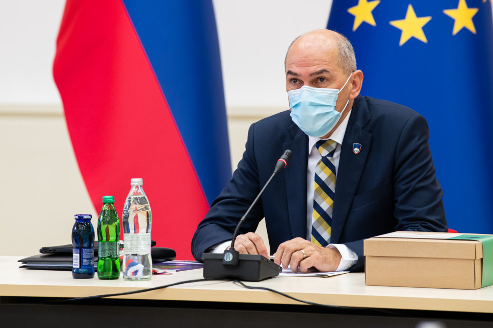 Janez Janša sedi za mizo, v ozadju so zastave.