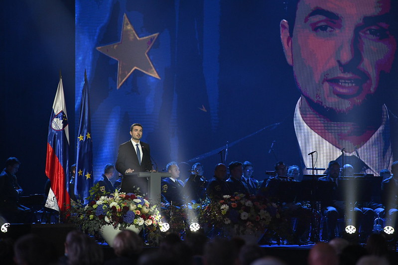 Minister Tonin za govornico in na zaslonu, v ozadju glasbeniki ter zastavi Slovenije in EU