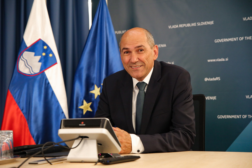 Janez Janša sedi za mizo, pred seboj ima računalnik, na levi so zastave.
