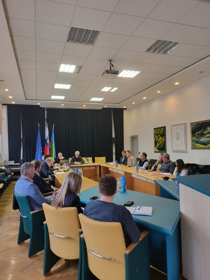 Moški in ženske sedijo za mizo in se pogovarjajo. Za njimi zastave.