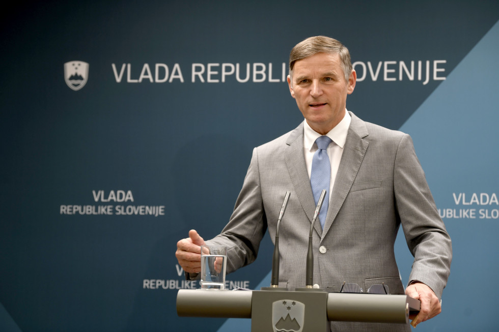 Minister stoji za govorniškim pultom in pojasnjuje na vladi sprejete odločitve. V ozadju je pano z napisom Vlada Republike Slovenije.