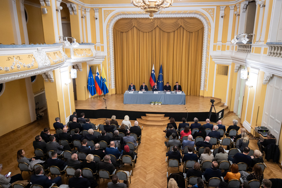 Dvorana, v kateri sedijo udeleženci. Na odru miza, za katero sedijo predstavniki vlade. Za njimi slovenska in evropska zastava.