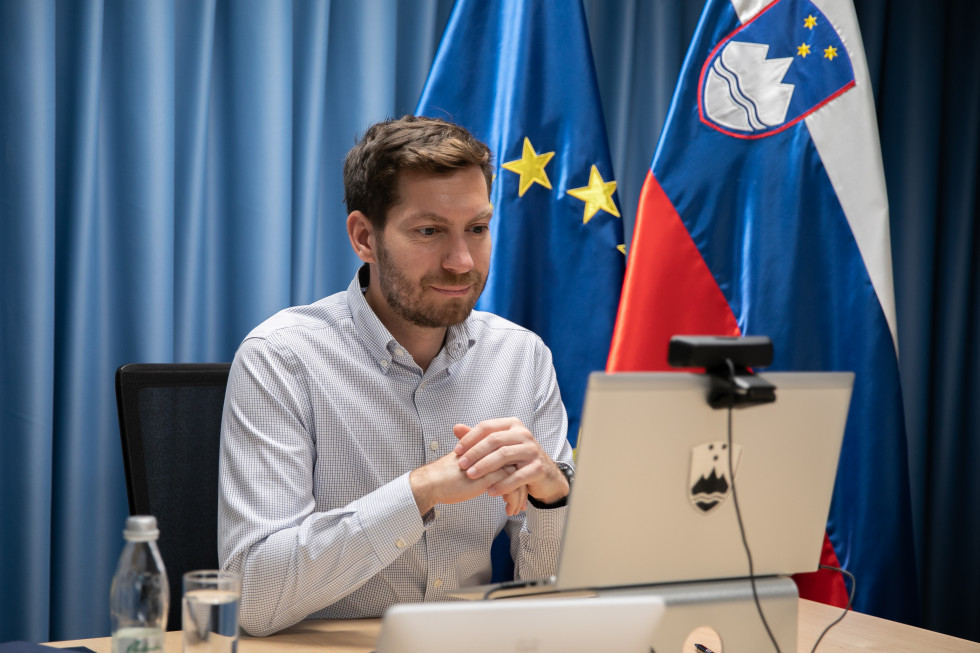 Mark Boris Andrijanič ob zaslonu računalnika, v ozadju zastavi Slovenije in EU