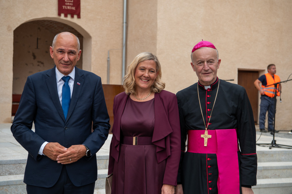 Moški in ženska stojijo ob katoliškem škofu, ki nosi vijolična liturgična oblačila in križ.