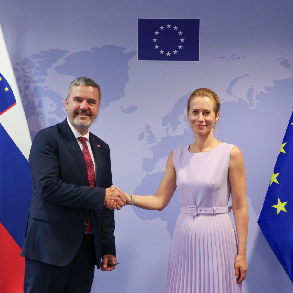 Two officials shake hands before an EU backdrop with flags; man in dark suit, red tie, woman in lavender dress.